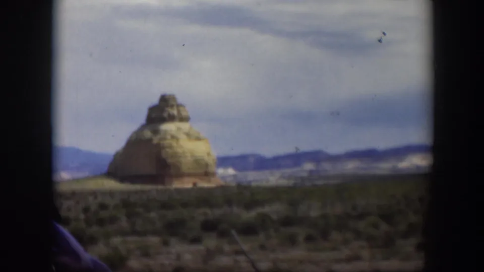1951 Utah road-trip frame showing a tan desert rock formation, low vegetation, hills, and dark vehicle framing at the sides.