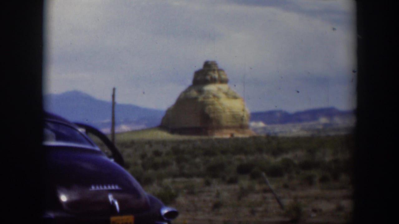 1951 Utah home-movie frame with a dark parked car in the foreground and a tan rock formation rising beyond scrub desert.