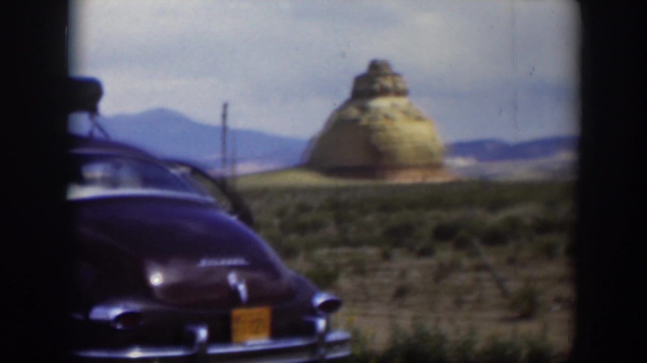 1951 Utah road-trip view of a dark parked car from the rear, with a tan rock formation, utility poles, and distant hills.