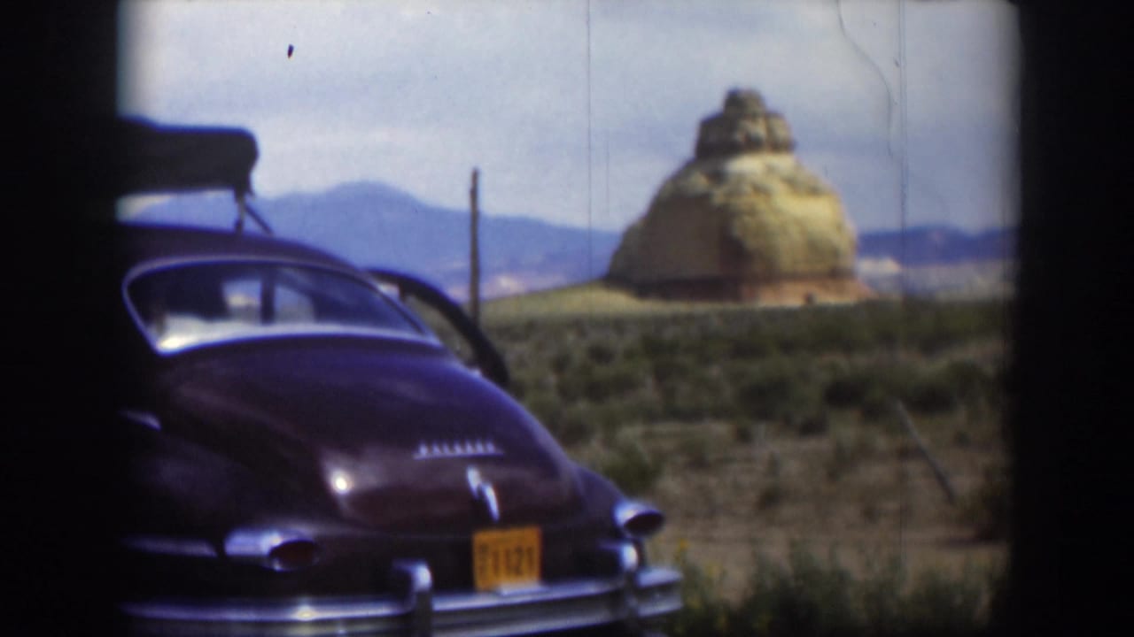Brown vintage sedan parked beside scrubby desert with a tan rock formation behind it.