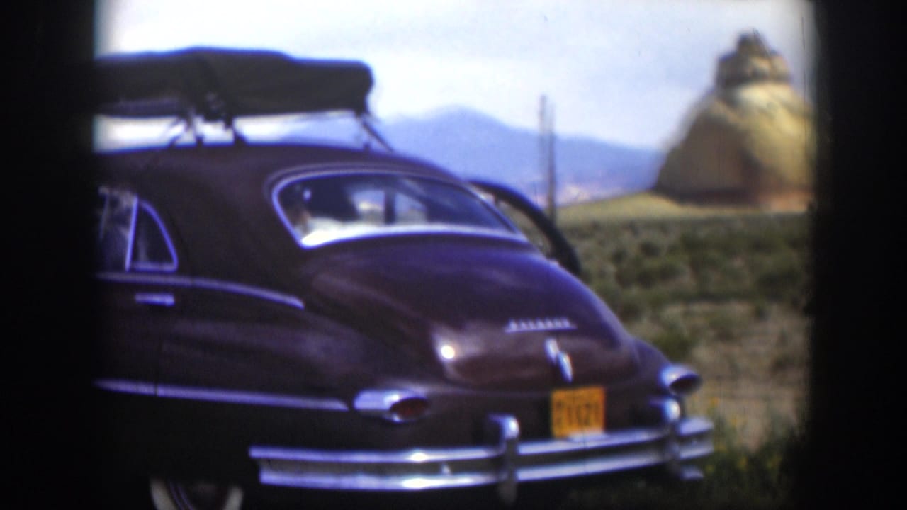 1951 Utah home-movie frame with a brown mid-century sedan, roof rack, yellow plate, scrubland, and tan rock formation in the background.