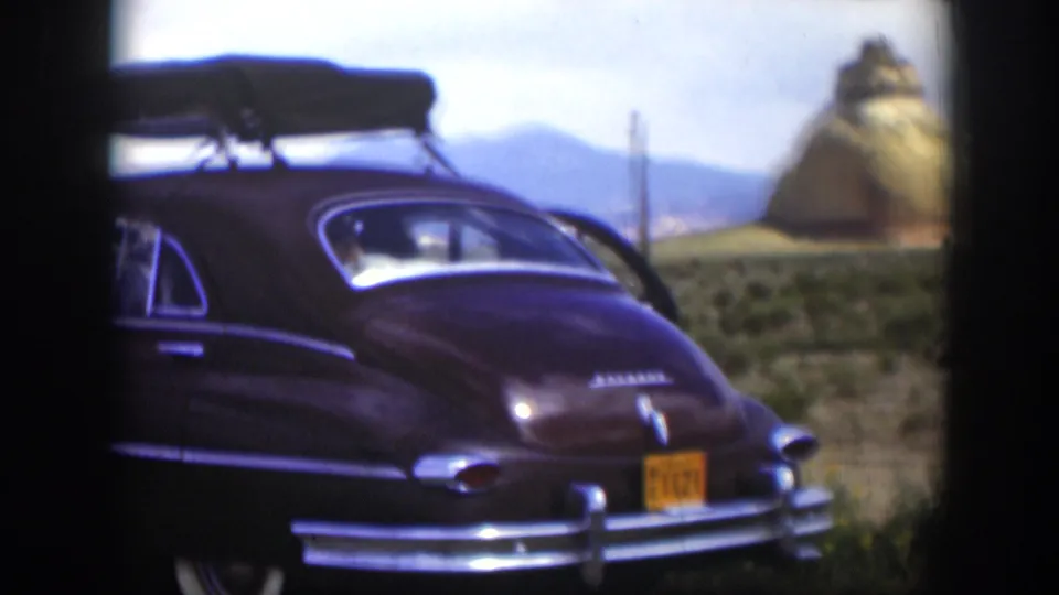 1951 Utah home-movie frame with a brown mid-century sedan, roof rack, yellow plate, scrubland, and tan rock formation in the background.