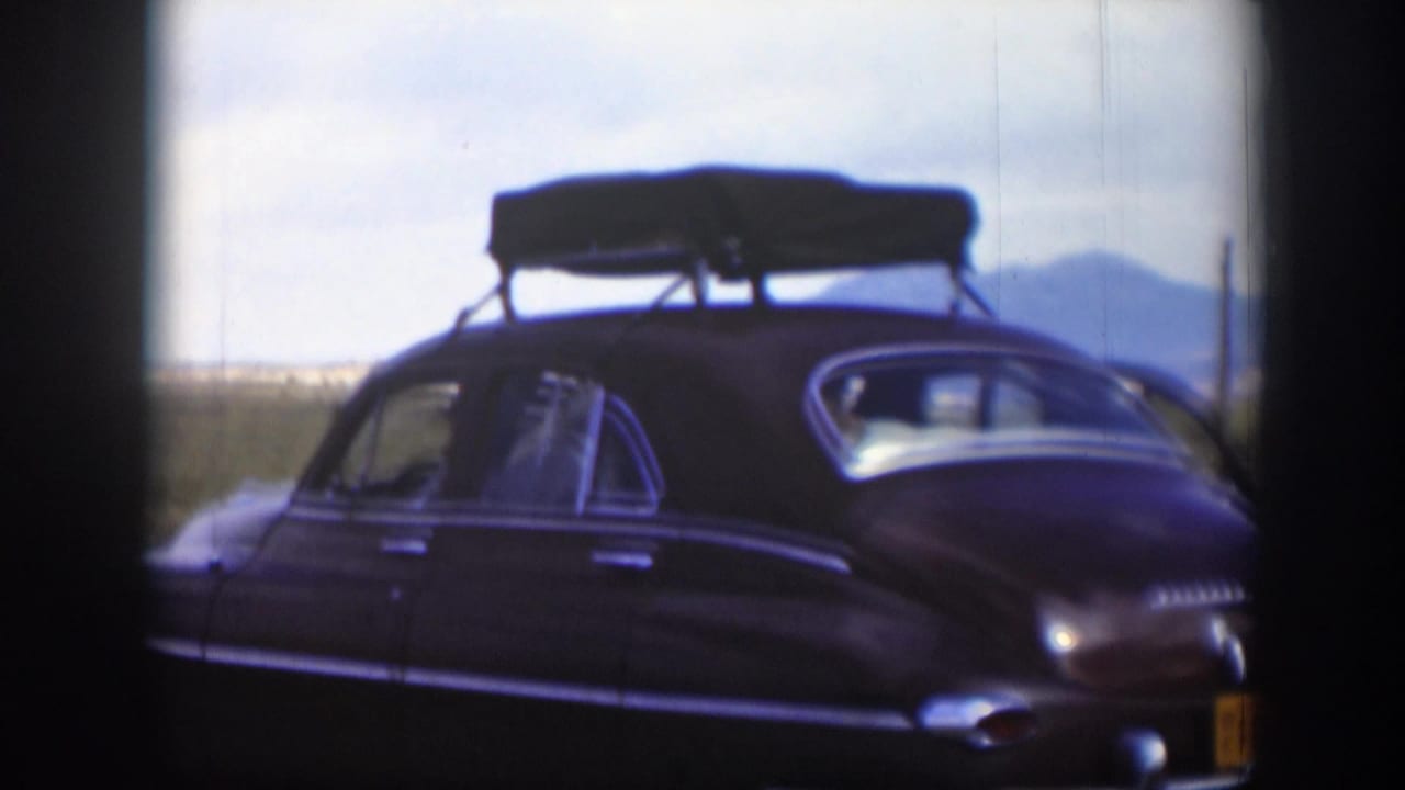 1951 Utah 8mm frame showing the side and rear of a brown sedan with roof luggage, desert brush, mountains, and soft film edges.
