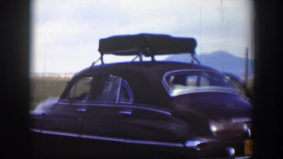 1951 Utah 8mm frame showing the side and rear of a brown sedan with roof luggage, desert brush, mountains, and soft film edges.