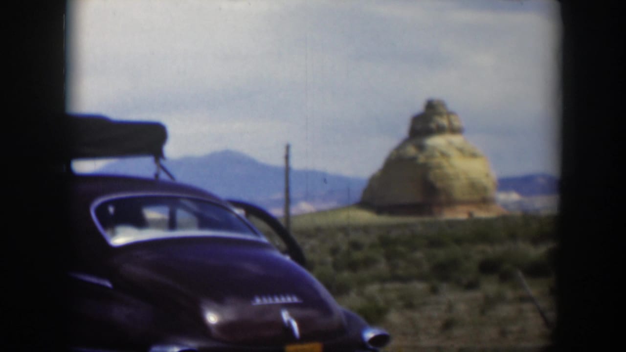 1951 Utah frame of a brown sedan seen from behind, with a tan rock formation, scrubland, distant mountains, and dark film-frame borders.