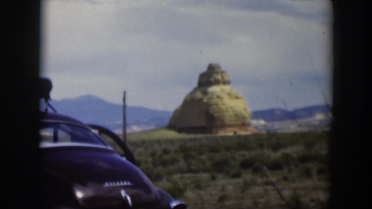Dark sedan edge beside a layered desert rock formation under a cloudy sky.