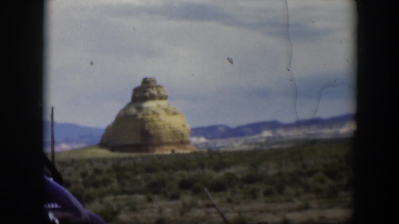 1951 Utah home-movie frame with a butte-like rock formation, arid foreground, low mountains, and the edge of a dark car.