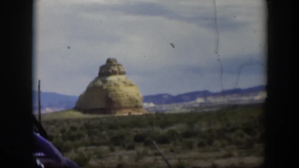 1951 Utah home-movie frame with a butte-like rock formation, arid foreground, low mountains, and the edge of a dark car.