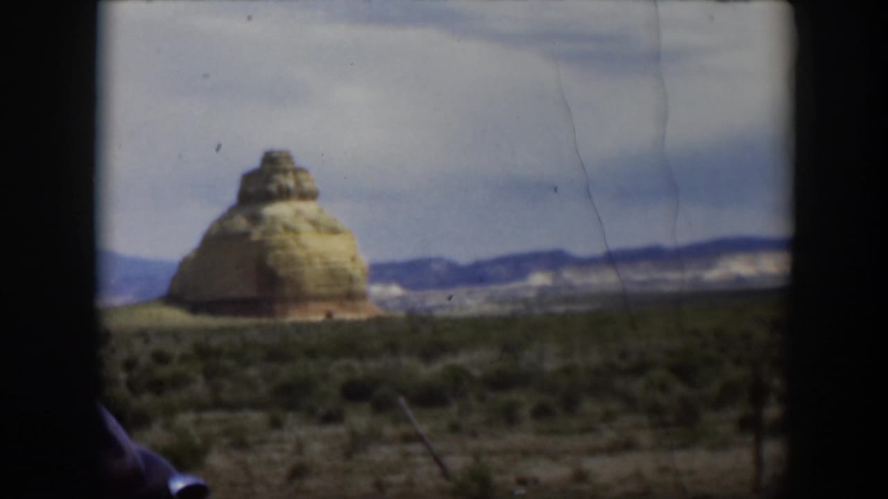 1951 Utah 8mm frame of a layered desert formation at left, open scrubland, distant mountains, and a partial car edge.