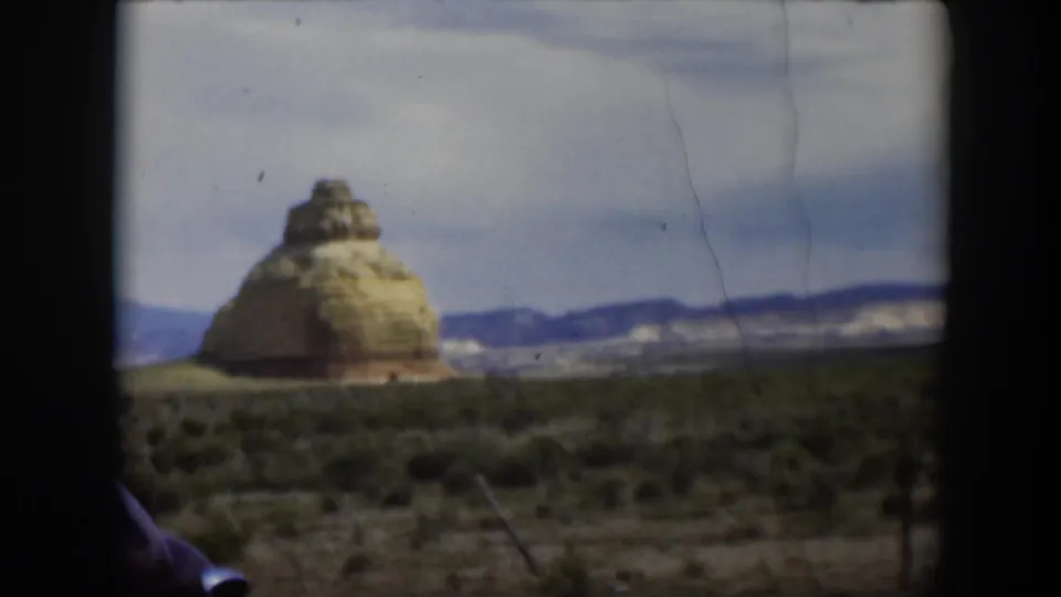 1951 Utah 8mm frame of a layered desert formation at left, open scrubland, distant mountains, and a partial car edge.