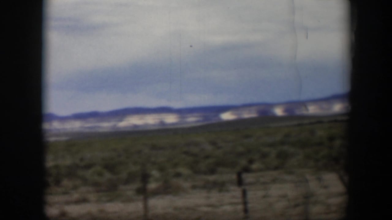 1951 Utah desert landscape with low shrubs, distant mountains, cloudy sky, and dark edges from filming inside or beside a vehicle.