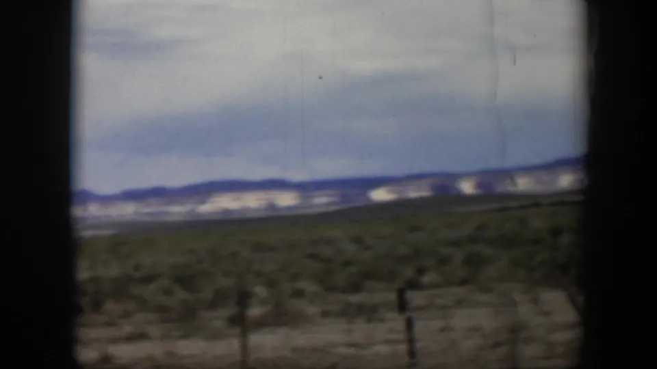 1951 Utah desert landscape with low shrubs, distant mountains, cloudy sky, and dark edges from filming inside or beside a vehicle.