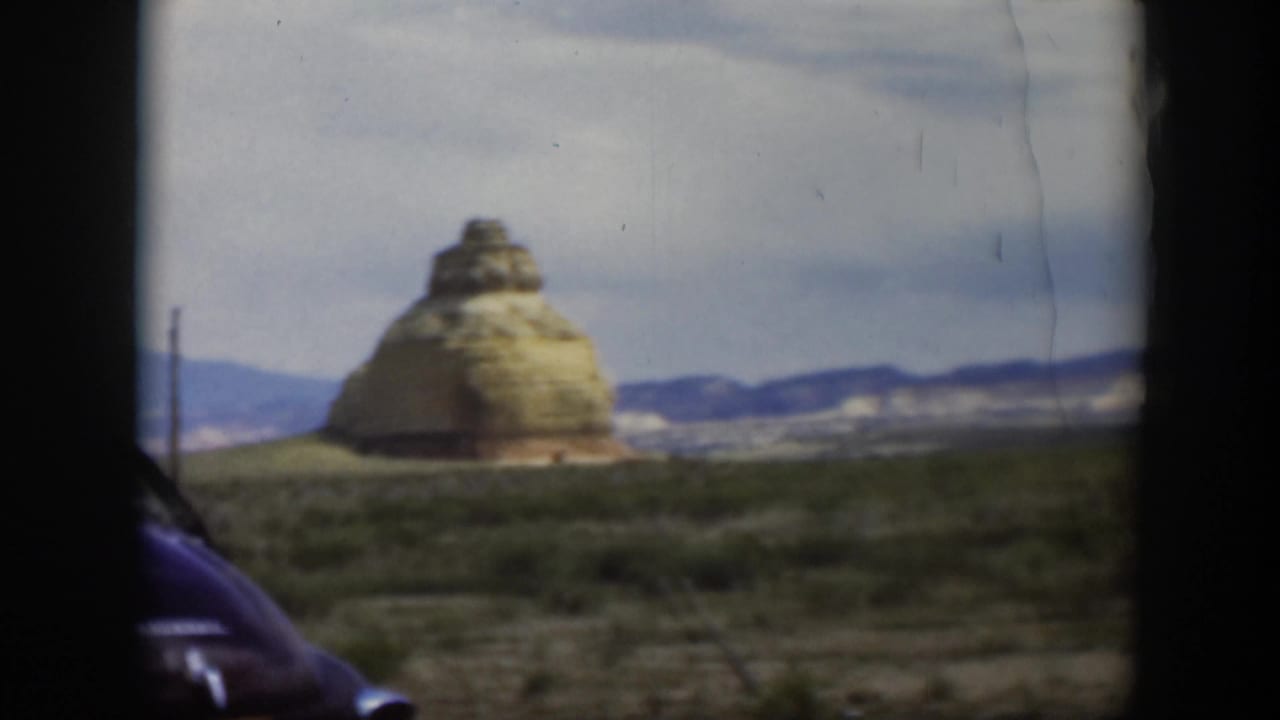 Distinctive rounded sandstone formation in a Utah desert view, with sparse brush, road-edge posts, and mountains in the distance.