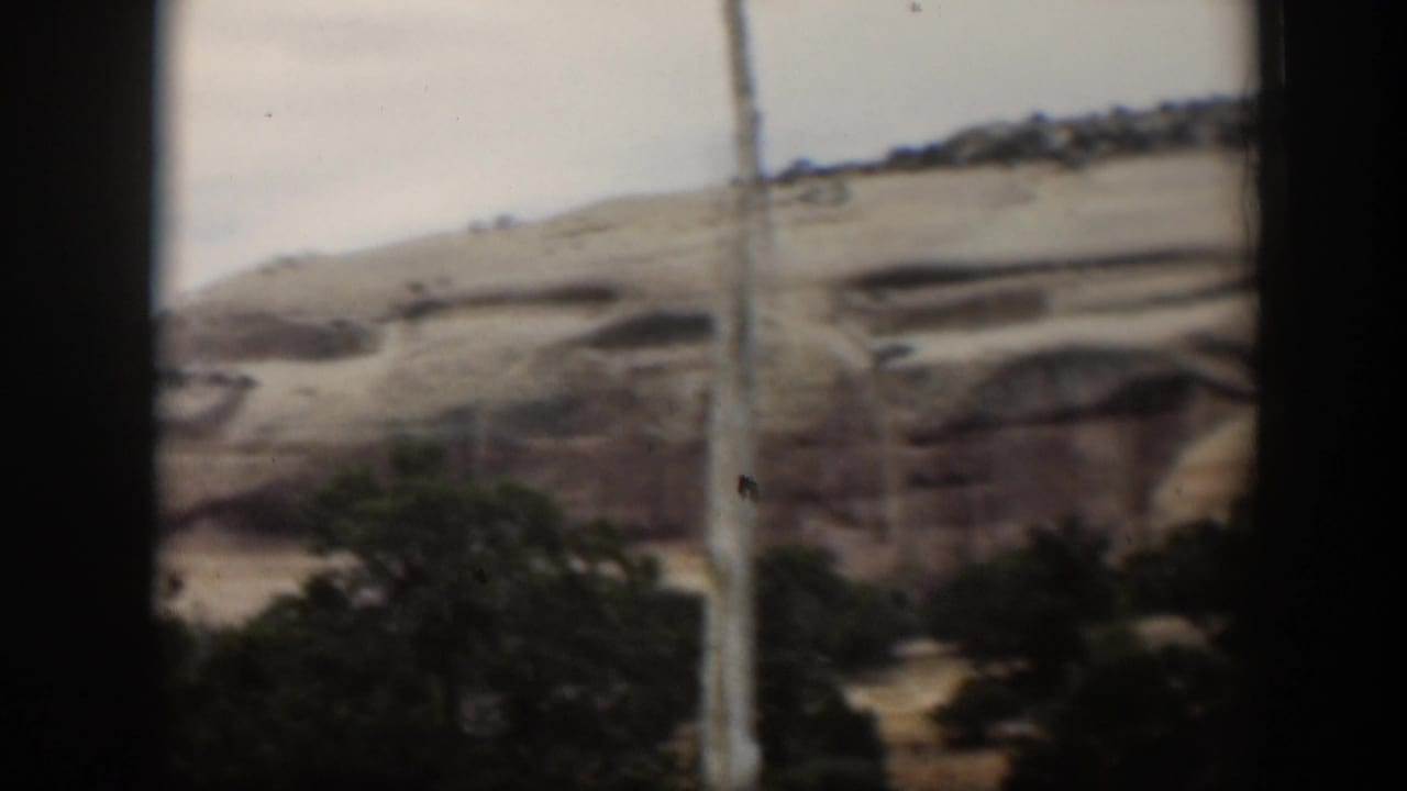 Utah sandstone cliffs and eroded pale rock faces pass by in soft-focus 8mm footage, with shrubs along the lower edge of frame.