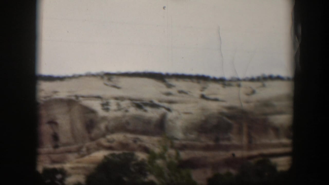 A broad Utah sandstone slope fills the frame, with sparse vegetation at the base and home-movie film marks visible over the pale sky.