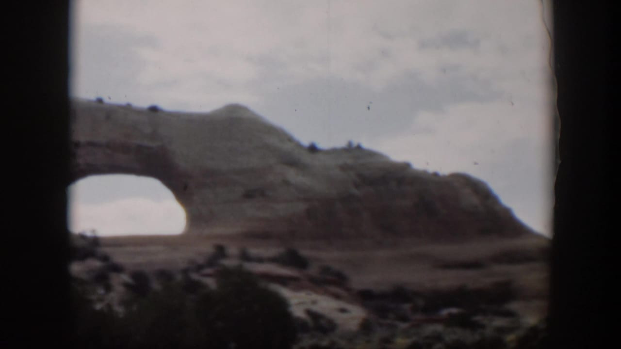 1951 Utah home-movie view of a large natural rock arch under a pale sky, with dark vehicle-window framing in the foreground.