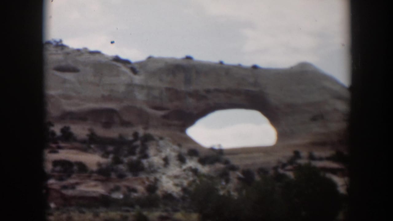 1951 Utah home-movie frame of a broad natural arch across a rocky desert scene, with shrubs and dark foreground borders visible.