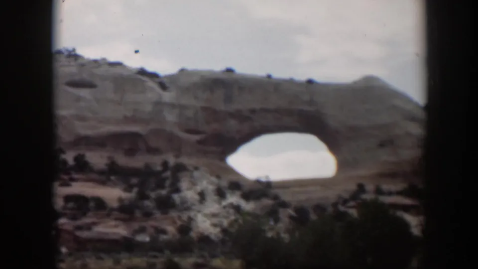 1951 Utah home-movie frame of a broad natural arch across a rocky desert scene, with shrubs and dark foreground borders visible.