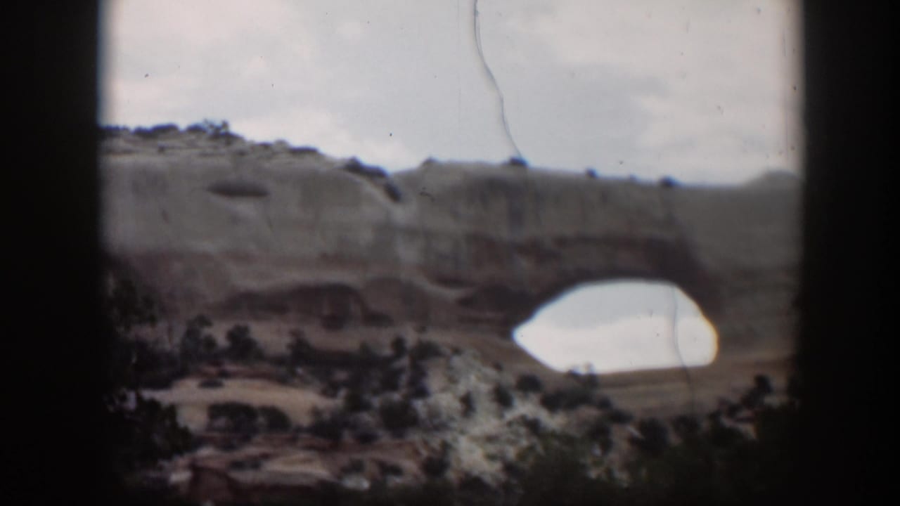 1951 Utah 8mm frame showing a tan sandstone arch and layered mesa with green shrubs below and cloudy sky above.