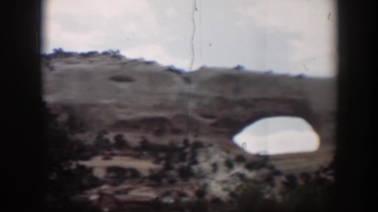 1951 Utah 8mm view of a natural sandstone arch in a mesa wall, with scrub vegetation and soft overcast light.