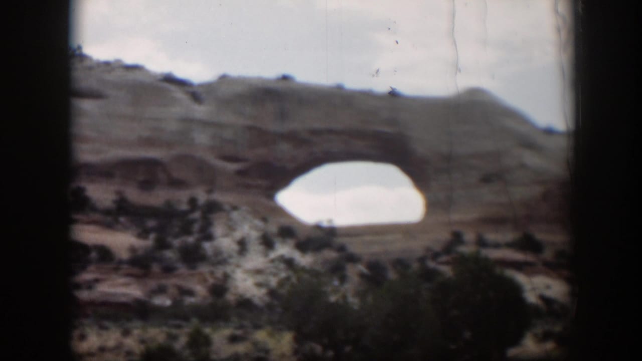 1951 Utah home-movie frame with a closer, soft-focus view of the tan rock arch above desert vegetation.