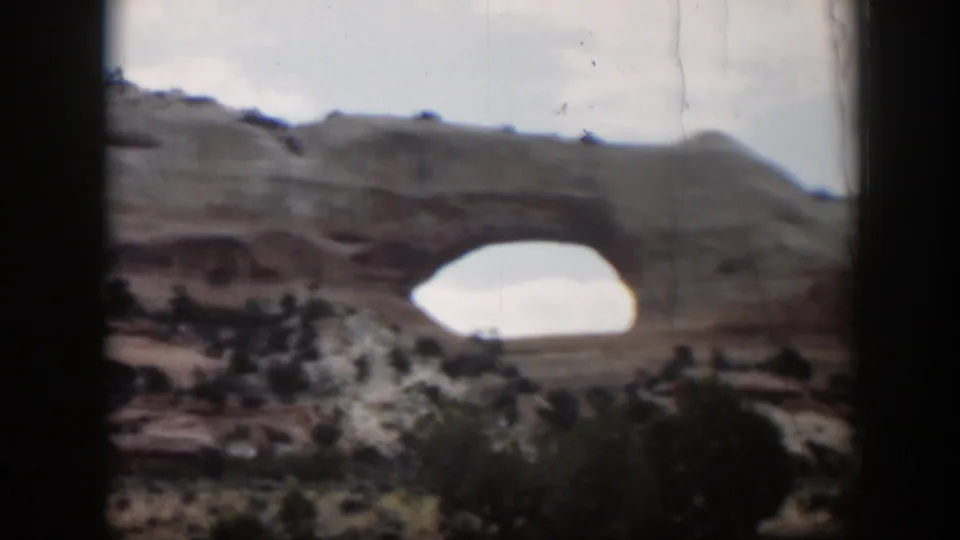 1951 Utah home-movie frame with a closer, soft-focus view of the tan rock arch above desert vegetation.