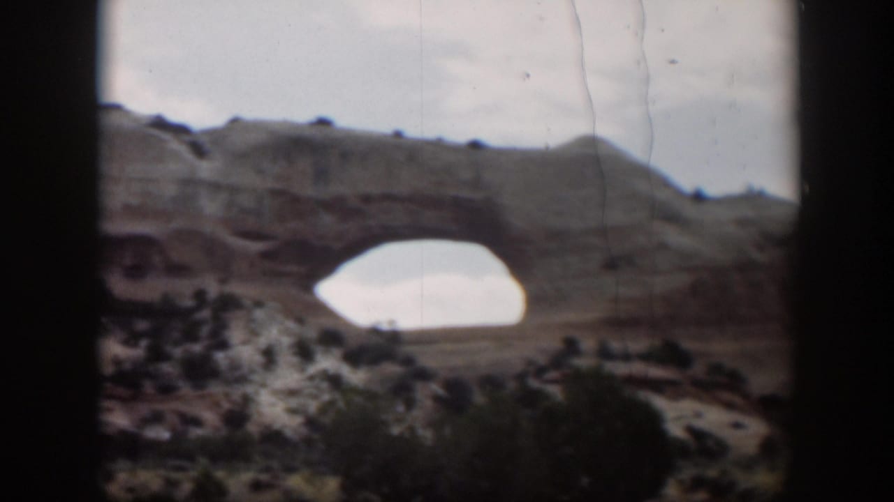 1951 Utah 8mm frame showing the sandstone arch from a steady visitor viewpoint, with cloudy sky and scrub vegetation.