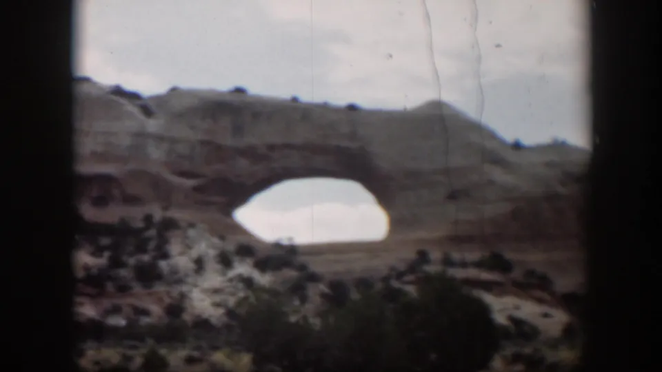 1951 Utah 8mm frame showing the sandstone arch from a steady visitor viewpoint, with cloudy sky and scrub vegetation.