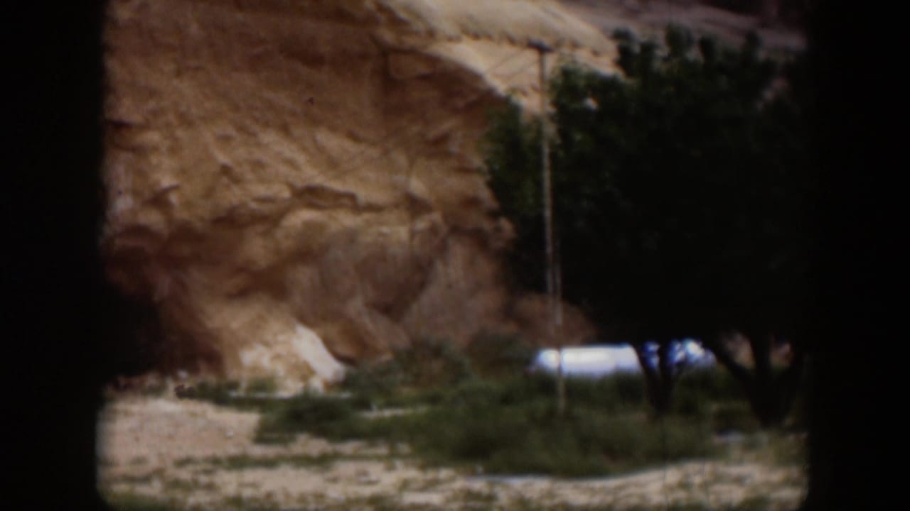1951 Utah desert frame with an eroded sandstone cliff, sparse vegetation, and a pale vehicle or trailer-like object partly visible near a tree.