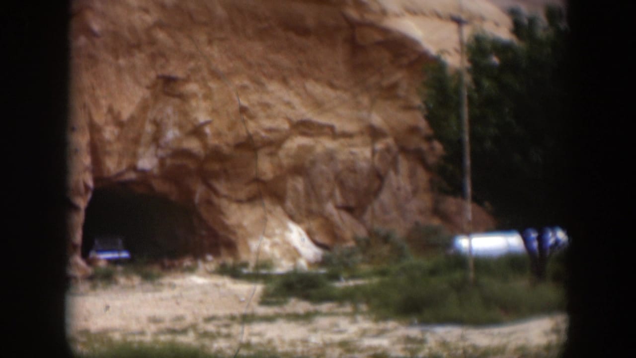 1951 Utah 8mm frame showing a dark cave entrance in a sandstone wall, with low shrubs and a white vehicle or trailer-like shape nearby.
