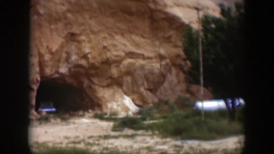 1951 Utah 8mm frame showing a dark cave entrance in a sandstone wall, with low shrubs and a white vehicle or trailer-like shape nearby.