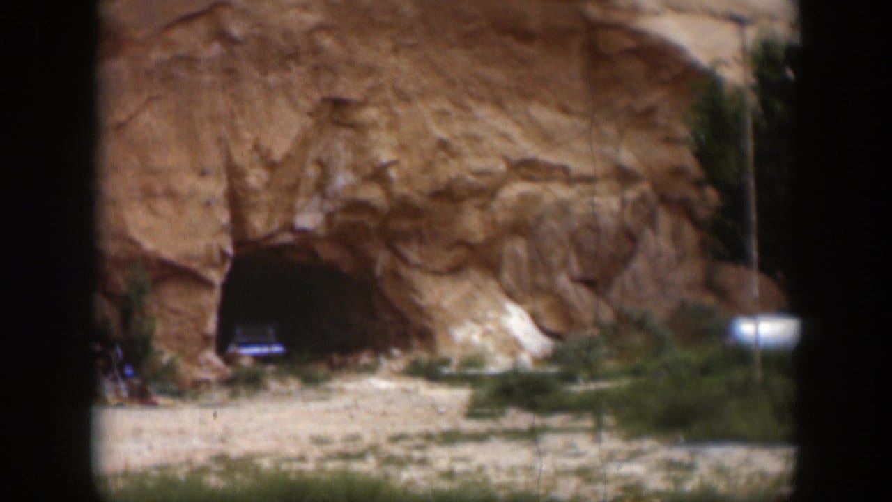 1951 Utah desert frame centered on a cliffside cave entrance, with a dark vehicle-like shape inside and sparse roadside vegetation.