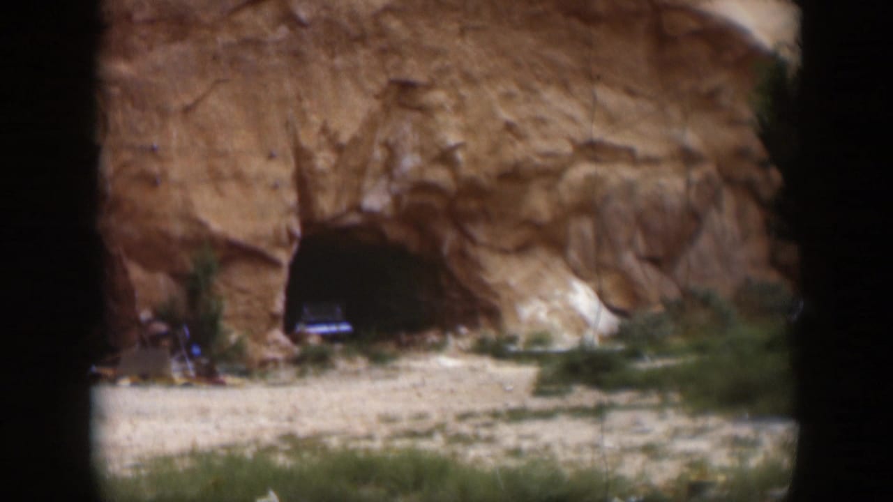 1951 Utah home-movie frame showing a sandstone rock face with two dark cave-like openings and sparse vegetation at the base.