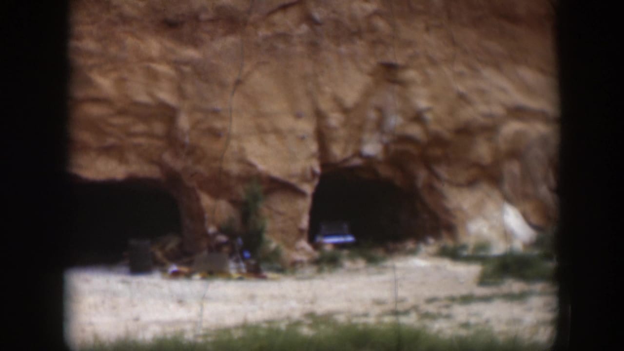 1951 Utah frame showing cave-like openings in a reddish rock face, with a parked car near the right opening.
