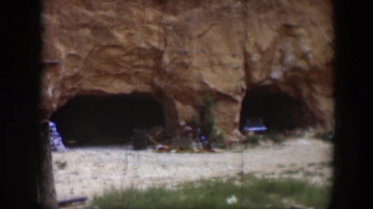 1951 Utah home-movie frame with a closer view of two shadowed openings in a red rock face and scattered items near the base.