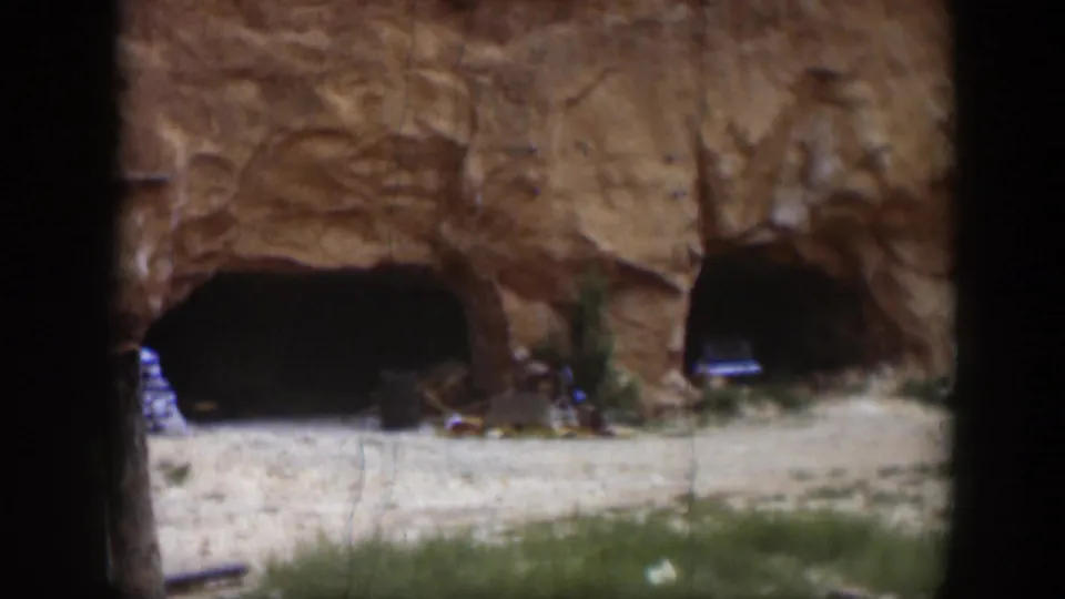1951 Utah home-movie frame with a closer view of two shadowed openings in a red rock face and scattered items near the base.