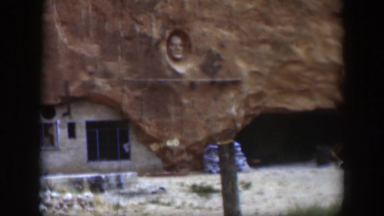 1951 Utah frame showing a small rock-built structure with windows and a doorway beneath a carved face in sandstone.