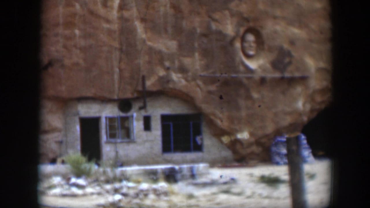 Sandstone cliff with a small building front, windows, open doorway, and a carved face above.