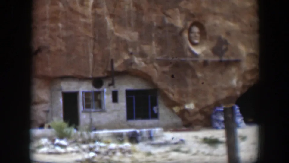 1951 Utah 8mm frame showing a small structure built into a sandstone rock face with windows, an open doorway, scrub plants, and a carved face above.