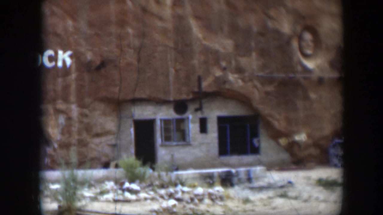 1951 Utah home-movie frame of a cliffside building beneath sandstone, with partial painted lettering, a window, a doorway, and a carved face above.