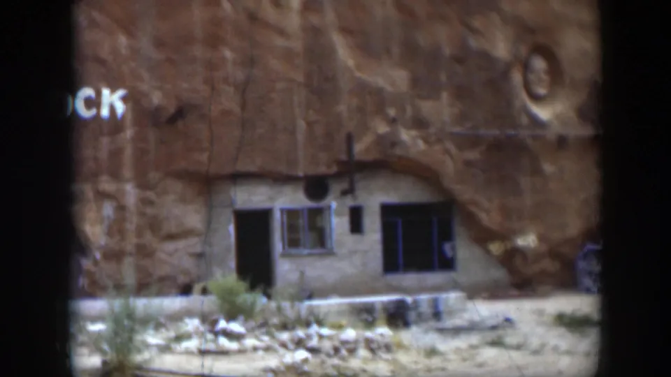 1951 Utah home-movie frame of a cliffside building beneath sandstone, with partial painted lettering, a window, a doorway, and a carved face above.