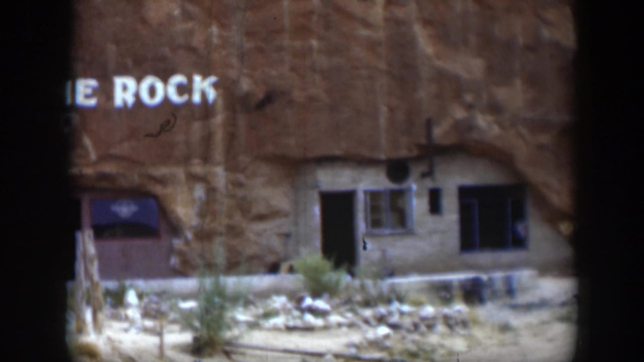 1951 Utah 8mm frame showing white painted words on sandstone above a modest rock-front building with a dark doorway, window, and dry ground.