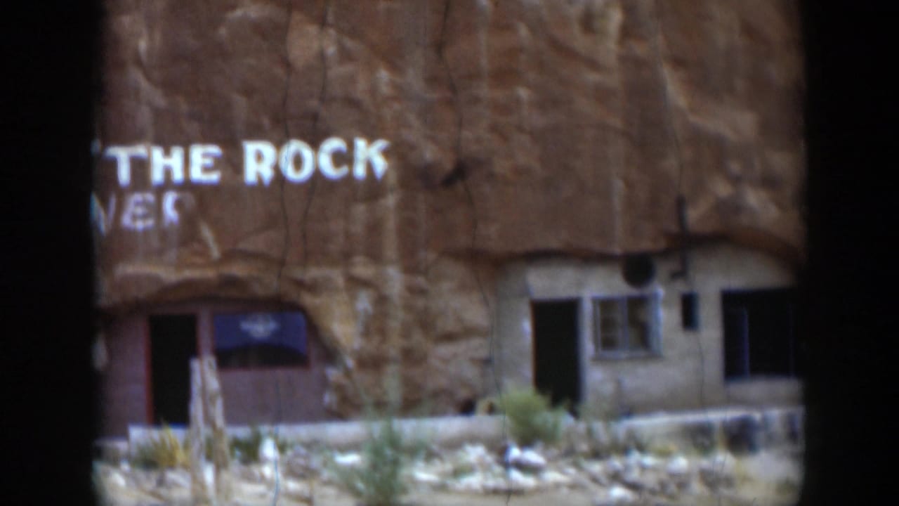 1951 Utah home-movie frame with visible THE ROCK lettering on a sandstone face, a small doorway and window below, and sparse desert vegetation in front.