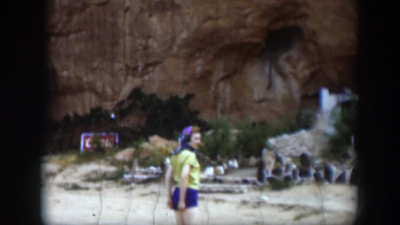 1951 Utah home-movie frame of a woman posing near the Hole N' The Rock setting, with sandstone cliff and desert ground behind her.