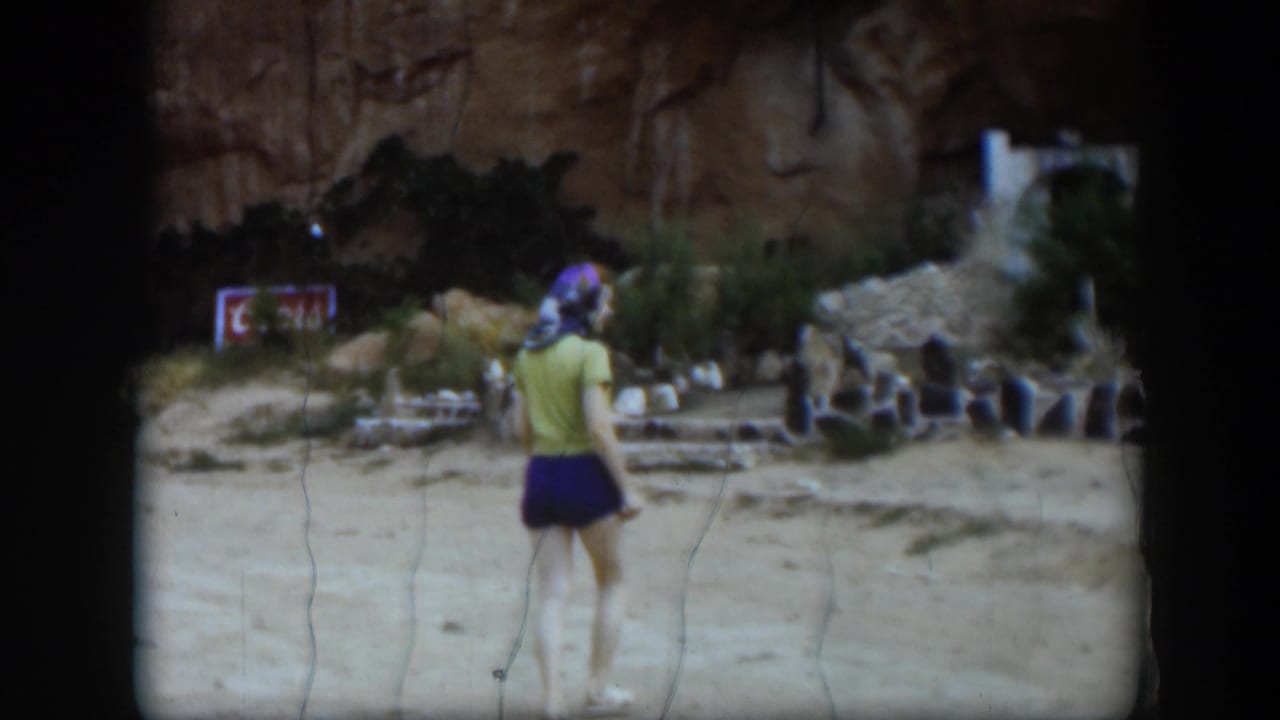 1951 Utah frame showing a woman from behind near the cliffside roadside attraction, with shrubs, stone borders, and sandstone visible.