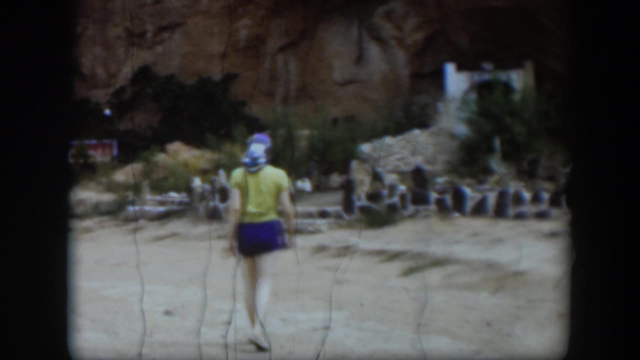 In 1951 Utah home-movie footage, a woman in a headscarf walks away from the camera across sandy ground toward stone ruins and a rock wall.