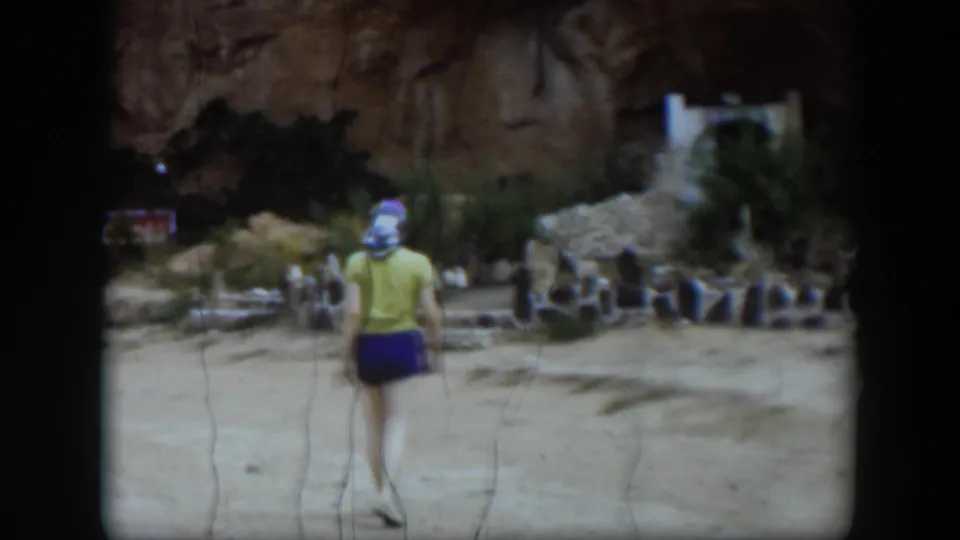 In 1951 Utah home-movie footage, a woman in a headscarf walks away from the camera across sandy ground toward stone ruins and a rock wall.