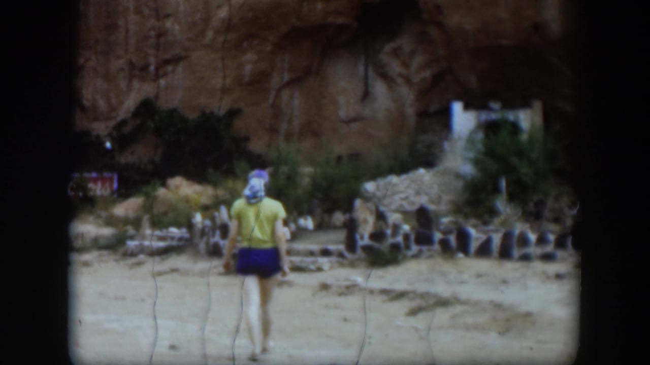 The woman continues across the dry Utah site, with low stone structures, brush, and a pale opening in the rocky background.