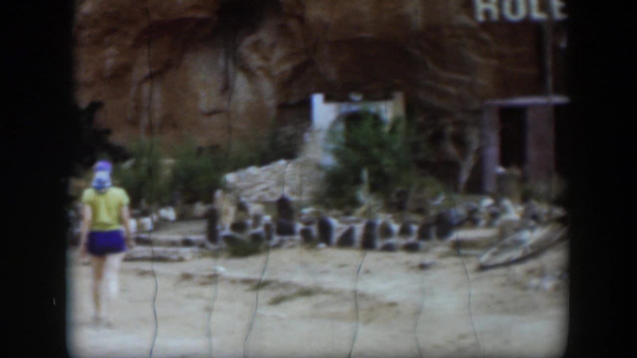 The 1951 Utah frame shows the woman moving out of view beside low ruins, dry ground, a rock wall, and part of a sign near a doorway-like structure.