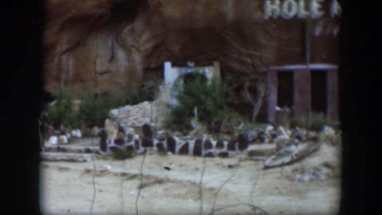 The final frame holds on the Utah ruin area, showing low stone remains, sandy ground, dry vegetation, a partial sign, and a small structure at right.